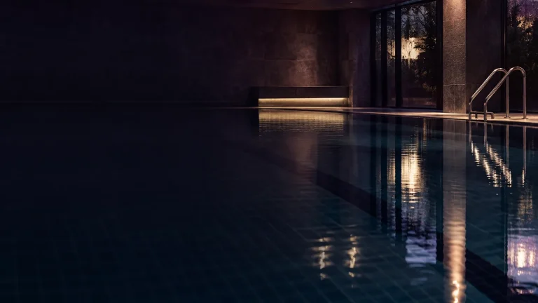A dark indoor swimming pool at night, its still water reflecting a few warm lights.