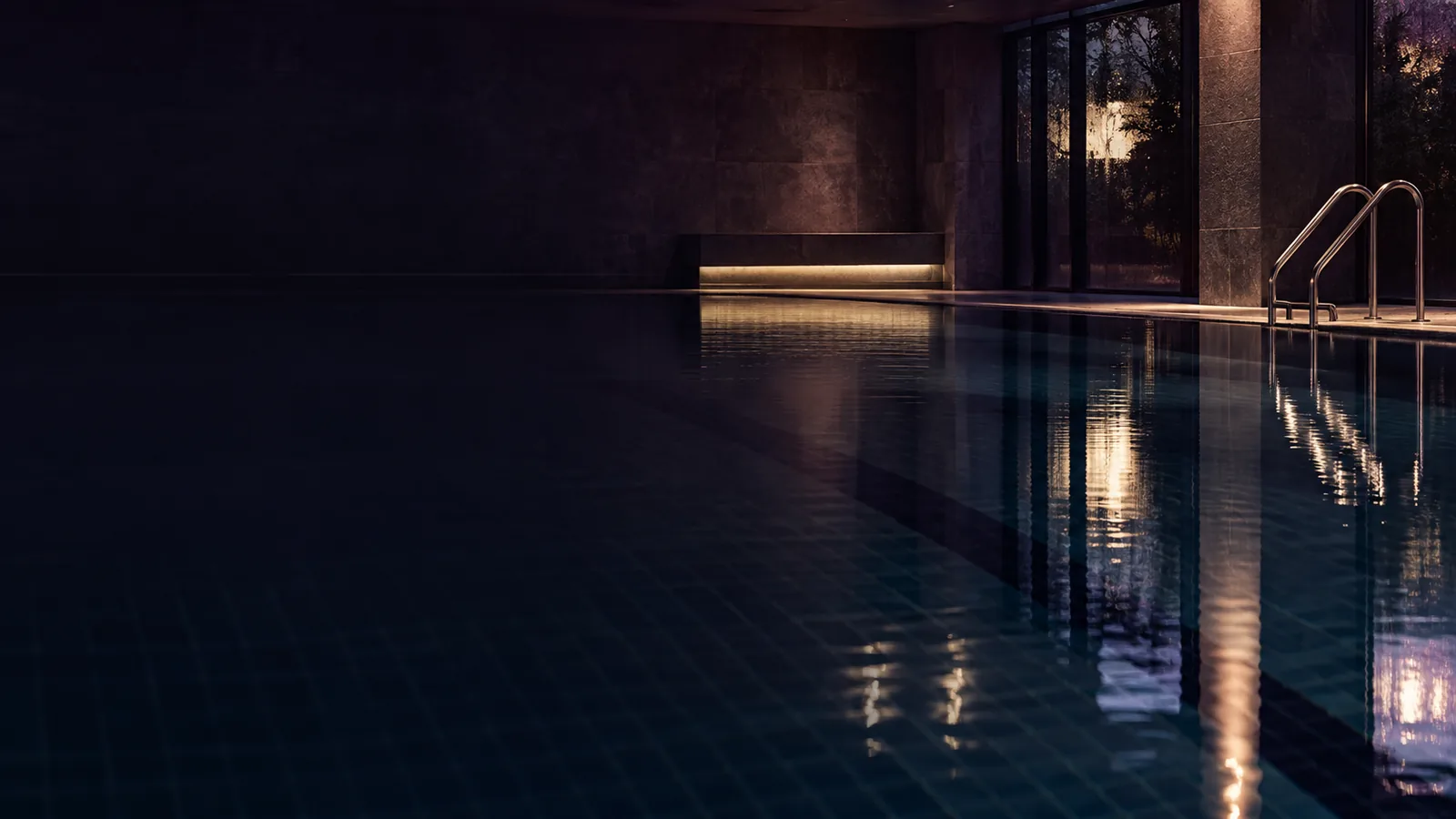 A dark indoor swimming pool at night, its still water reflecting a few warm lights.